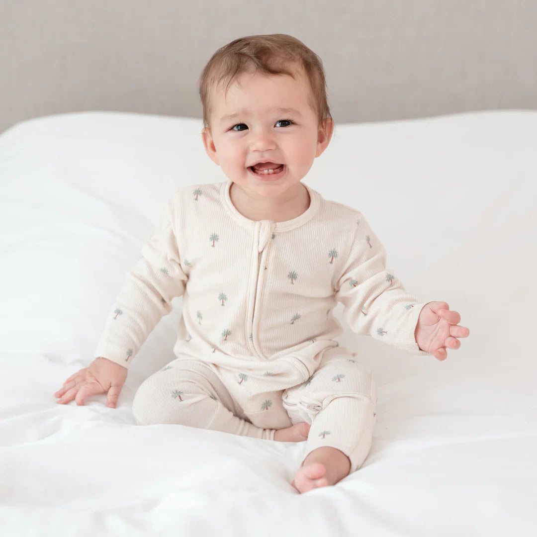 Baby sitting on a white blanket wearing a cream-colored sleepsuit with date palm from Hushabye from The Conscious Collective 
