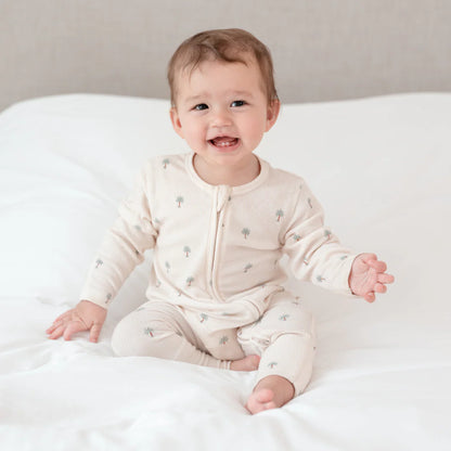 Baby sitting on a white blanket wearing a cream-colored sleepsuit with date palm from Hushabye from The Conscious Collective 